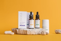 A collection of skincare products is displayed on a textured stone platform against a vibrant orange background. The items include two brown dropper bottles labeled 'Premium CBD Oil' and a white box labeled 'Acne Treatment CBD Cream.' Additional bottles and textured stones are arranged artistically around the platform.