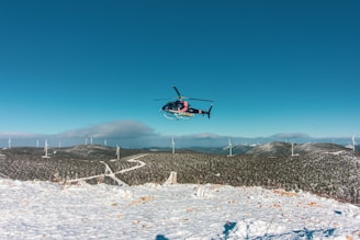 A helicopter is flying above a snowy landscape dotted with multiple wind turbines. The clear blue sky stretches expansively above, while low-lying clouds hover near the horizon. Snow covers the ground and the hills in the background.