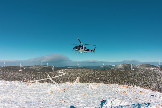 A helicopter is flying above a snowy landscape dotted with multiple wind turbines. The clear blue sky stretches expansively above, while low-lying clouds hover near the horizon. Snow covers the ground and the hills in the background.