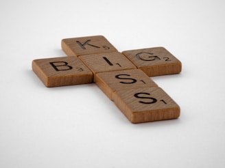 Wooden Scrabble tiles arranged on a white surface to form the words 'BIG' and 'KISS'. The tiles are light brown with black letters and point values.