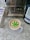 A cup of coffee with a latte art design resembling a cannabis leaf in green, set on a gray saucer. To the left, there is a carton of oat milk with text and branding visible. On the right, a package of CBD infused coffee is partially visible. The background consists of a coffee machine with a metallic surface and a stone countertop.