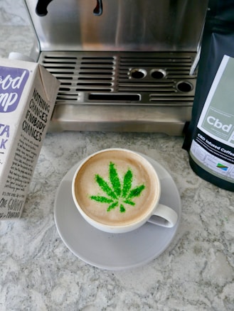 A cup of coffee with a latte art design resembling a cannabis leaf in green, set on a gray saucer. To the left, there is a carton of oat milk with text and branding visible. On the right, a package of CBD infused coffee is partially visible. The background consists of a coffee machine with a metallic surface and a stone countertop.