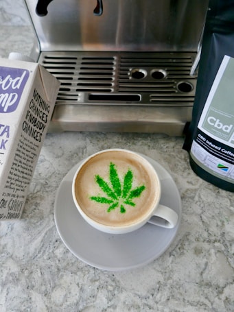A cup of coffee with a latte art design resembling a cannabis leaf in green, set on a gray saucer. To the left, there is a carton of oat milk with text and branding visible. On the right, a package of CBD infused coffee is partially visible. The background consists of a coffee machine with a metallic surface and a stone countertop.