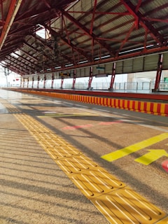 An empty railway platform with a slanted red roof and exposed steel beams, featuring a line of tactile paving on the ground. The sunlight creates distinct shadows, and there are directional arrows in bright yellow and red on the platform floor.