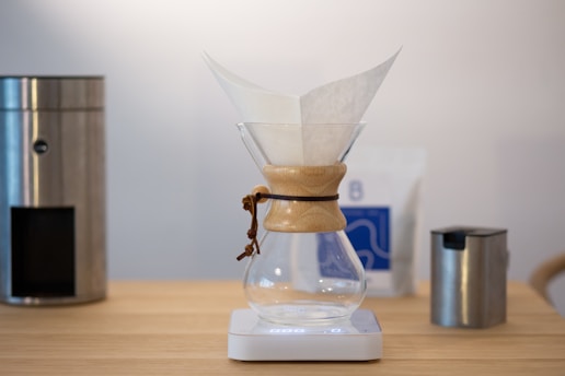 A modern kitchen setup featuring a Chemex coffee maker with a filter paper inside, placed on a digital scale. Surrounding it are various metal kitchen appliances, including a coffee grinder and a milk frother, all on a wooden countertop.