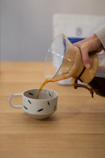 A cozy scene of a hand pouring freshly brewed coffee from a classic pour-over dripper into a warm ceramic cup on a wooden table.