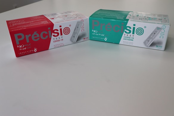 Two boxes of COVID-19 test kits are placed on a flat surface. The box on the left is predominantly red, while the box on the right is primarily green. Both boxes have the brand name 'Precisio' prominently displayed, along with text indicating they are for COVID-19 testing.