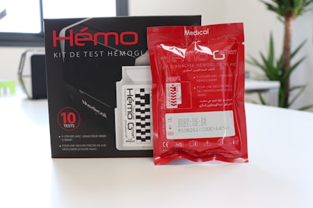 A hemoglobin testing kit with a box containing multiple test kits. The packaging includes detailed labeling in both French and Arabic, with a red foil packet placed in front of the box. The text provides product information, usage instructions, and expiration dates.