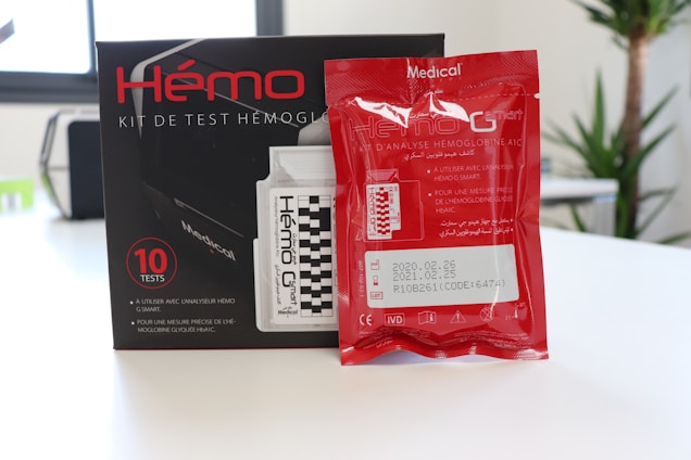 A hemoglobin testing kit with a box containing multiple test kits. The packaging includes detailed labeling in both French and Arabic, with a red foil packet placed in front of the box. The text provides product information, usage instructions, and expiration dates.