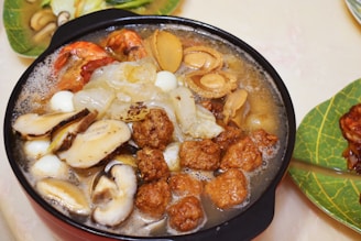 A sizzling hotpot filled with a variety of ingredients including shrimp, mushrooms, quail eggs, meatballs, and abalone. The dish is rich in colors and textures, steaming hot and ready to serve. The background shows a leafy serving plate with additional food items.