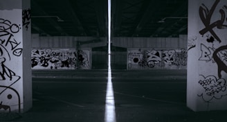 A dimly lit urban underpass with multiple concrete pillars, each covered in various graffiti tags and artwork. The area appears desolate and quiet, with a narrow beam of light cutting through the center, creating a stark contrast against the dark surroundings.