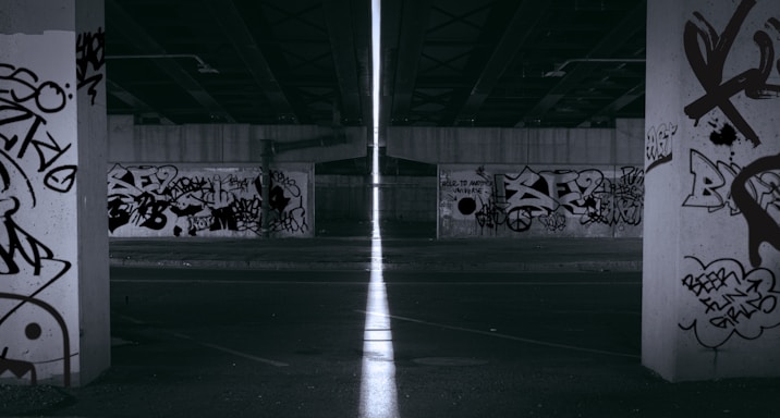 A dimly lit urban underpass with multiple concrete pillars, each covered in various graffiti tags and artwork. The area appears desolate and quiet, with a narrow beam of light cutting through the center, creating a stark contrast against the dark surroundings.