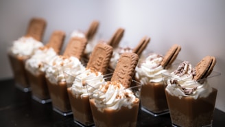 A row of desserts in clear cups, each featuring a layer of creamy pudding topped with whipped cream, a biscuit, and chocolate pieces. The presentation is neat and appealing, with the desserts aligned on a dark surface.