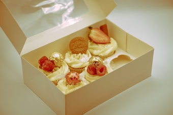 A box of assorted cupcakes with various toppings sits open. The cupcakes are decorated with whipped cream and garnished with raspberries, strawberries, chocolates, and a cookie with a brand imprint. The box is positioned on a flat surface, and the cupcakes display intricate detailing.
