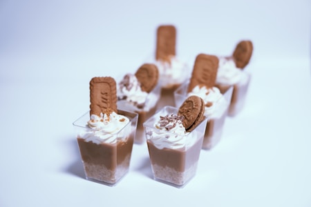 A series of small dessert cups, each filled with layers of whipped cream, chocolate mousse, and crunchy biscuit. The servings are topped with cookies and some chocolate shavings, placed in a neat arrangement on a light surface.