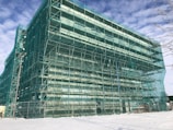 A building covered with green scaffolding, indicating active construction or renovation work. The structure is large and surrounded by a snowy landscape under a partly cloudy sky.