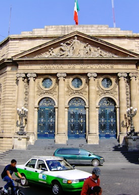 A grand neoclassical building facade with ornate columns and detailed carvings above the entrance. The building is labeled 'Camara de Diputados' with a Mexican flag flying at the top. In front, there is a green and white taxi, a person on a bicycle, and another individual walking, suggesting an urban setting.