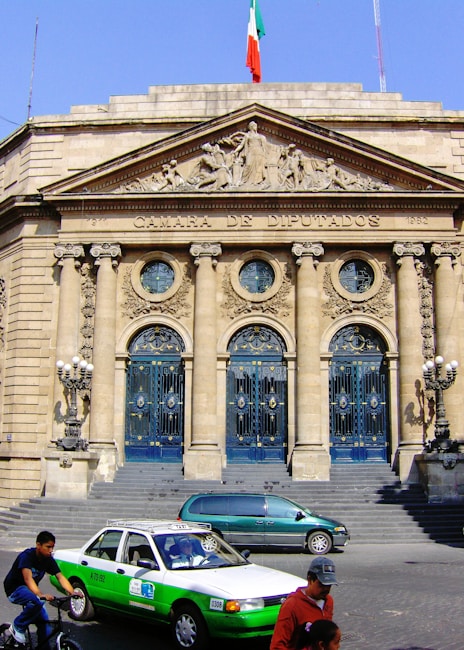 A grand neoclassical building facade with ornate columns and detailed carvings above the entrance. The building is labeled 'Camara de Diputados' with a Mexican flag flying at the top. In front, there is a green and white taxi, a person on a bicycle, and another individual walking, suggesting an urban setting.