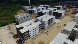 Aerial view of a construction site featuring multiple new, multi-story apartment buildings. Each building has a similar design with a combination of white and dark grey exteriors. The area is surrounded by greenery and suburban residential buildings in the background.