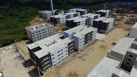 Aerial view of a construction site featuring multiple new, multi-story apartment buildings. Each building has a similar design with a combination of white and dark grey exteriors. The area is surrounded by greenery and suburban residential buildings in the background.