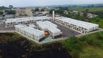 Aerial view of a residential complex with uniform white-roofed buildings arranged in a three-sided layout. The structures are surrounded by a mix of urban and semi-rural greenery, with houses and trees visible in the background. The complex is bordered by a grassy area and has a central asphalt parking lot.