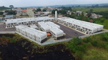 Aerial view of a residential complex with uniform white-roofed buildings arranged in a three-sided layout. The structures are surrounded by a mix of urban and semi-rural greenery, with houses and trees visible in the background. The complex is bordered by a grassy area and has a central asphalt parking lot.