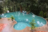 A uniquely shaped, turquoise swimming pool surrounded by lush green trees and foliage. The pool appears to have a tree integrated into its design, growing in the center of one of its sections. The poolside is bordered with tan tiles, and various potted plants decorate the area, complementing the natural scenery. The atmosphere is serene and tranquil, offering a harmonious blend of nature and leisure.