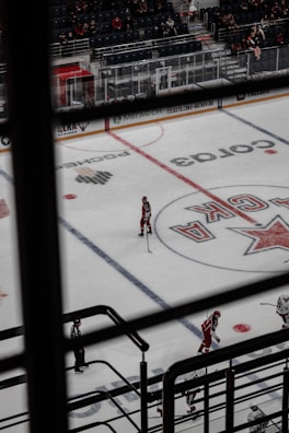 An indoor hockey game is taking place, viewed from above through a set of railings. The ice rink displays markings and logos, with several players dressed in red uniforms and one dressed in striped referee attire. The arena's seating area is partially filled with spectators.