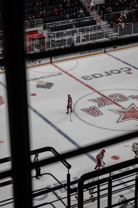 An indoor hockey game is taking place, viewed from above through a set of railings. The ice rink displays markings and logos, with several players dressed in red uniforms and one dressed in striped referee attire. The arena's seating area is partially filled with spectators.