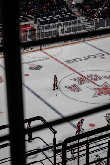 An indoor hockey game is taking place, viewed from above through a set of railings. The ice rink displays markings and logos, with several players dressed in red uniforms and one dressed in striped referee attire. The arena's seating area is partially filled with spectators.
