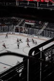 A professional ice hockey game is captured from an elevated perspective in an indoor arena. Players from two teams are actively engaged in play, with some clustered near the goal. The ice rink is surrounded by clear plexiglass barriers and there are advertisements on the boards. Spectators are seated in the background, and the seating area has red and blue colors. The arena features a prominent lighting system illuminating the ice.
