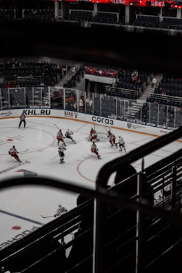 A professional ice hockey game is captured from an elevated perspective in an indoor arena. Players from two teams are actively engaged in play, with some clustered near the goal. The ice rink is surrounded by clear plexiglass barriers and there are advertisements on the boards. Spectators are seated in the background, and the seating area has red and blue colors. The arena features a prominent lighting system illuminating the ice.
