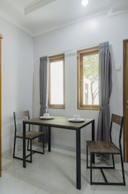A small dining room with a modern minimalist design features a rectangular table with a dark wooden top and black metal legs. Two matching chairs are placed on either side of the table, which is set with white cups and saucers. The room is brightly lit by natural light coming through two windows with gray curtains, creating a clean and cozy atmosphere.