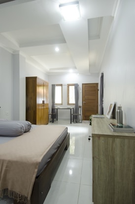 A modern, minimalist bedroom with a neatly made bed featuring soft pillows and a beige blanket. The room has a wooden wardrobe and a small desk with two chairs near the window. The color palette is neutral, with light walls and a polished white tile floor. A small side table holds a water bottle and a few books, while soft lighting from the ceiling brightens the space.