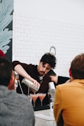 A bartender is skillfully pouring drinks from a shaker into cocktail glasses. The bar setting includes several people observing, with a tiled wall and decorative artwork in the background.