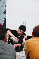 A bartender is skillfully pouring drinks from a shaker into cocktail glasses. The bar setting includes several people observing, with a tiled wall and decorative artwork in the background.