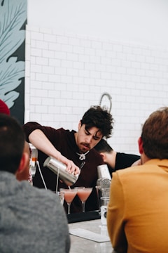 A bartender is skillfully pouring drinks from a shaker into cocktail glasses. The bar setting includes several people observing, with a tiled wall and decorative artwork in the background.