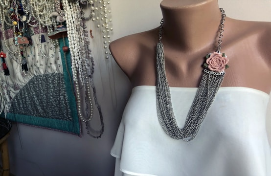 A mannequin torso is adorned with a chain necklace featuring a pink rose embellishment. The mannequin wears a white top. In the background, there is a display of various necklaces and beads hanging on the wall.