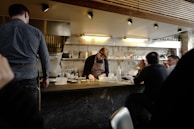 A chef working behind a counter in a small, cozy restaurant setting. Several patrons are seated at the bar, watching the chef as he prepares food. The space has a warm, rustic feel with exposed stone walls and wooden paneling on the ceiling. Shelves with glasses, bottles, and cooking utensils line the wall behind the chef.