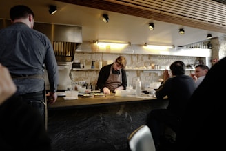 A chef working behind a counter in a small, cozy restaurant setting. Several patrons are seated at the bar, watching the chef as he prepares food. The space has a warm, rustic feel with exposed stone walls and wooden paneling on the ceiling. Shelves with glasses, bottles, and cooking utensils line the wall behind the chef.