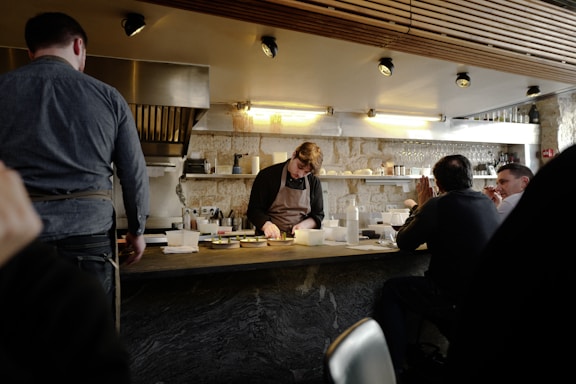A chef working behind a counter in a small, cozy restaurant setting. Several patrons are seated at the bar, watching the chef as he prepares food. The space has a warm, rustic feel with exposed stone walls and wooden paneling on the ceiling. Shelves with glasses, bottles, and cooking utensils line the wall behind the chef.