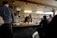 A chef working behind a counter in a small, cozy restaurant setting. Several patrons are seated at the bar, watching the chef as he prepares food. The space has a warm, rustic feel with exposed stone walls and wooden paneling on the ceiling. Shelves with glasses, bottles, and cooking utensils line the wall behind the chef.