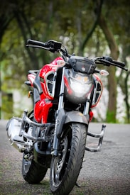 A sleek and modern red motorcycle with twin headlights parked on a road surrounded by trees. The design is sporty, featuring a prominent black front fender and handlebars. The background is a blurred natural setting, giving emphasis to the motorcycle's bright color and design.