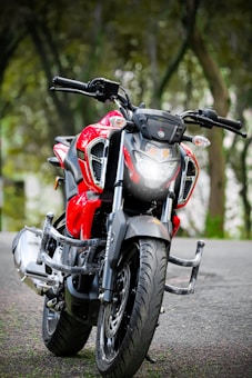 A sleek and modern red motorcycle with twin headlights parked on a road surrounded by trees. The design is sporty, featuring a prominent black front fender and handlebars. The background is a blurred natural setting, giving emphasis to the motorcycle's bright color and design.