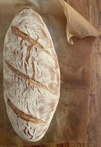 A rustic loaf of bread with a crusty, flour-dusted surface rests on a piece of brown parchment paper. The bread has a golden-brown color with distinct slashing marks across the top, indicating an artisanal baking style. The parchment paper is placed on a wooden surface.