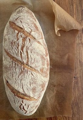 A rustic loaf of bread with a crusty, flour-dusted surface rests on a piece of brown parchment paper. The bread has a golden-brown color with distinct slashing marks across the top, indicating an artisanal baking style. The parchment paper is placed on a wooden surface.