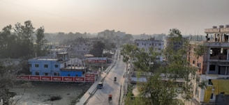 A semi-urban landscape featuring residential buildings, a wide road running through the center, and a few scattered vehicles. Trees and foliage are visible, adding greenery to the surroundings, while some construction work is noticeable on some buildings. The sky is slightly overcast, suggesting an early morning or late afternoon setting.