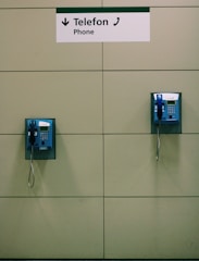 Two blue public payphones are mounted on a beige tiled wall, with a sign above them labeled 'Telefon' and 'Phone'.