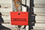 A red card with the word 'COMPASSION...' written in large black letters is attached to a metal latch on a weathered, rusted door.