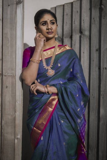 A woman in traditional Indian attire stands against a wooden backdrop. She is dressed in a rich blue and pink silk saree adorned with golden patterns. Her jewelry includes a heavy gold necklace, earrings, and bangles, along with a nose ring that adds to the traditional look. Her hair is neatly tied back, and she has a thoughtful expression.